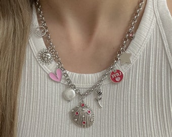 May include: A silver chain necklace with multiple charms, including a pink heart, a sun, a star, a pearl, a rose, and a seashell with red jewels. The charms are attached to a silver chain.