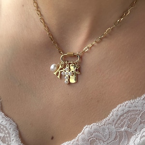 Op de afbeelding: Een gouden ketting met een bedel met een parel, een kruis, een kleine ster en een label met de letter "M".