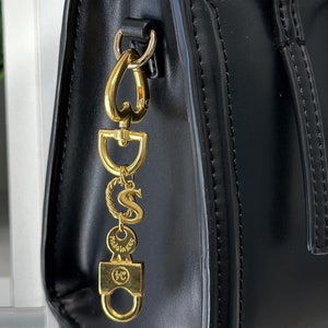 Può includere: Borsa nera con hardware color oro e un ciondolo decorativo. Il ciondolo presenta le lettere "D", "S" e "S", insieme ad altri elementi decorativi. La borsa ha una forma strutturata e un manico superiore.