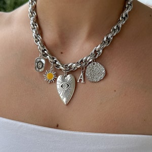 Puede incluir: Collar de cadena de plata con múltiples dijes, incluyendo un corazón con un ojo, un sol, una letra A y un círculo texturizado.