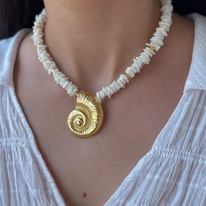 Può includere: Un pendente color oro a forma di conchiglia Nautilus è appeso a una collana di piccole perle bianche di forma irregolare. La collana è indossata con una camicetta bianca.