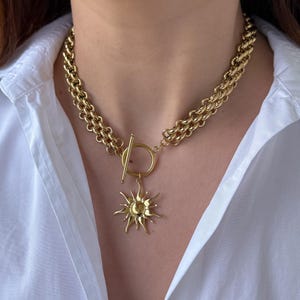 May include: A gold chain necklace with a large sun pendant. The chain is double-layered and has a toggle clasp.