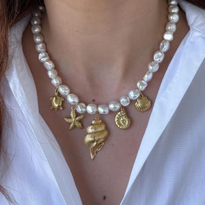 Puede incluir: Un collar de perlas blancas con colgantes de conchas y estrellas de mar doradas. El collar está puesto en una modelo que lleva una camisa blanca.