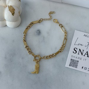 May include: Gold chain bracelet with a small gold boot charm. The bracelet is on a white surface with a white box and a white candle with gold accents in the background. The text "Love it SNAP IT SHARE IT" is on a white card with a gold star and a QR code.