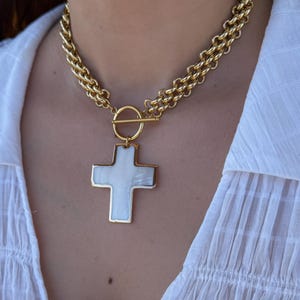 Peut inclure: Un collier chaîne doré avec un grand pendentif en forme de croix blanche. Le collier présente une chaîne à double rang et un fermoir à bascule. Le pendentif en croix a une bordure dorée et un centre blanc.