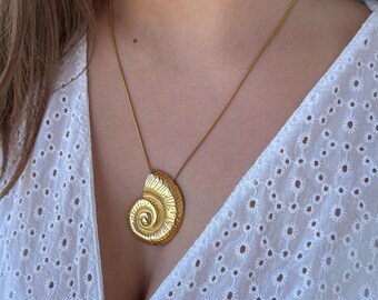 May include: A gold-colored pendant necklace featuring a spiral seashell design. The pendant hangs from a delicate gold chain. The necklace is worn over a white eyelet top.