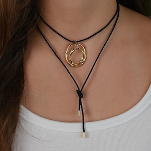 Può includere: Un girocollo nero con un ciondolo dorato composto da due cerchi intrecciati. La collana è regolabile con un nodo scorrevole.