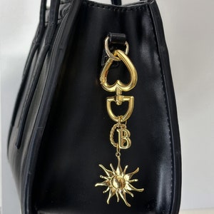 Puede incluir: Bolso tote de cuero negro con una cadena y un dije de oro con un corazón, la letra B y un diseño de sol.