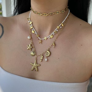 Puede incluir: Un collar de oro de varias capas con una variedad de dijes, incluyendo conchas marinas, estrellas de mar y un caballito de mar. El collar está hecho de cuentas blancas y eslabones de cadena de oro.