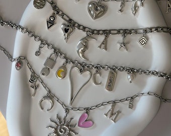 May include: A set of silver charm necklaces with various charms, including hearts, moons, stars, letters, and a sun with a spiral design. One charm is a pink heart.