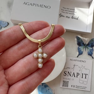 May include: Gold chain necklace with a small cross pendant adorned with white pearls. The necklace is displayed on a hand and comes in a white box with the text "AGAPIMENO" and "You are smart. You are beautiful. You are worthy. You are loved."