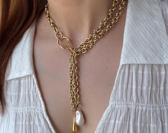 May include: Gold-toned necklace featuring a layered chain design. The necklace has a large ring detail and a dangling pendant with a pearl and a teardrop-shaped charm. The jewelry is worn over a white blouse.