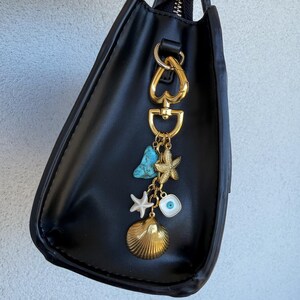 Può includere: Borsa nera con chiusura a forma di cuore color oro e un ciondolo con pietra turchese, stella marina, conchiglia e un ciondolo a forma di occhio. Il ciondolo è attaccato alla borsa con una catena color oro.
