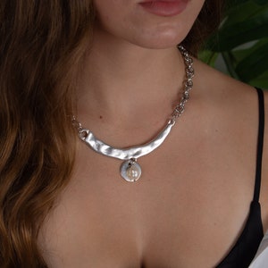 Könnte beinhalten: Eine silberne Halskette mit einem gehämmerten Metallanhänger und einem Perlenanhänger.
