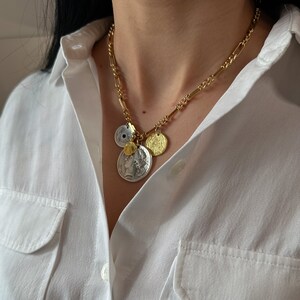 Puede incluir: Un collar de cadena dorada con m&uacute;ltiples colgantes en forma de moneda. Los colgantes son una mezcla de plata y oro, y algunos tienen una superficie texturizada.
