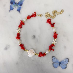 May include: A red coral and white pearl necklace with a seashell charm and starfish accents. The necklace is on a white marble surface.