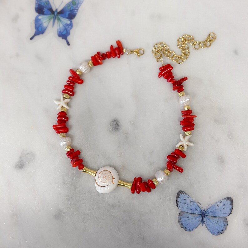 May include: A red coral and white pearl necklace with a seashell charm and starfish accents. The necklace is on a white marble surface.