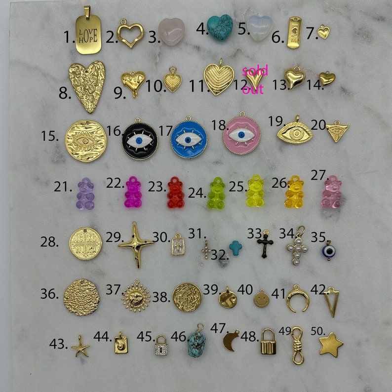 May include: Assortment of 50 gold-tone charms, including hearts, stars, crosses, eyes, bears, and other shapes. The charms are arranged in rows on a white surface, numbered 1 through 50.