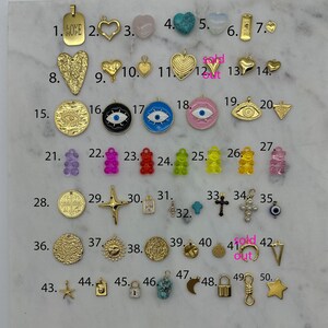May include: Assortment of 50 gold, silver, and colourful charms, including hearts, stars, crosses, eyes, bears, and more. The charms are numbered 1 through 50.
