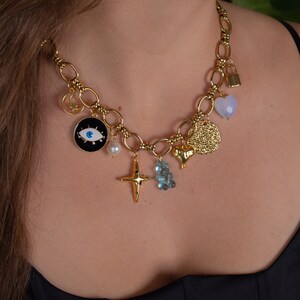 May include: A gold chain necklace with multiple charms, including a heart, a padlock, a cross, a pearl, a blue eye, and a gold coin. The charms are attached to a thick gold chain.