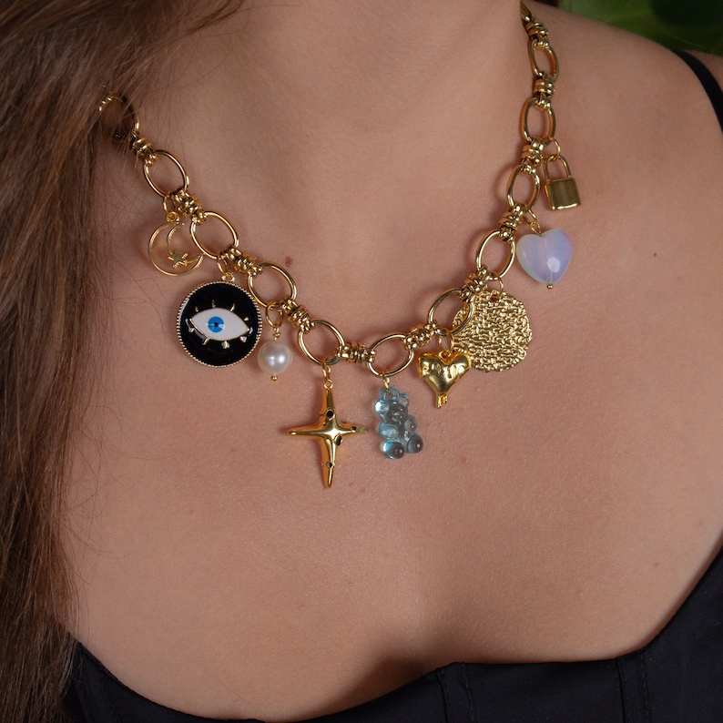 May include: A gold chain necklace with multiple charms, including a heart, a padlock, a cross, a pearl, a blue eye, and a gold coin. The charms are attached to a thick gold chain.