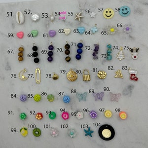 May include: Assortment of colourful charms and beads, including a seashell, a heart, a star, a smiley face, a flower, a Christmas tree, a stocking, a butterfly, a lemon, a kiwi, and a starfish. The charms are arranged in rows on a white surface.