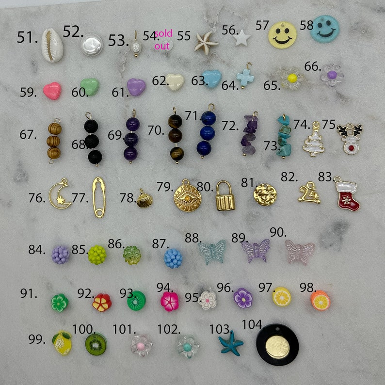May include: Assortment of colourful charms and beads, including a seashell, a heart, a star, a smiley face, a flower, a Christmas tree, a stocking, a butterfly, a lemon, a kiwi, and a starfish. The charms are arranged in rows on a white surface.