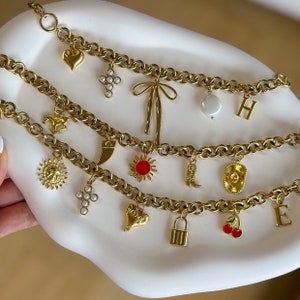 May include: A gold chain necklace with multiple charms, including a heart, a cross, a sun, a boot, a cowboy hat, a lock, and a cherry. The charms are all gold and have different designs.