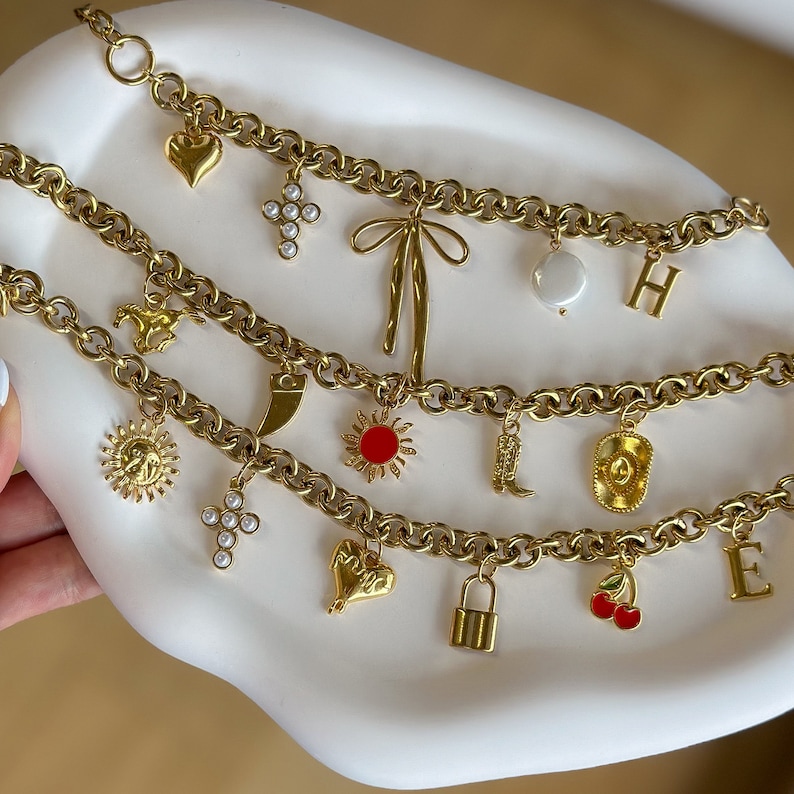 May include: A gold chain necklace with multiple charms, including a heart, a cross, a sun, a boot, a cowboy hat, a lock, and a cherry. The charms are all gold and have different designs.