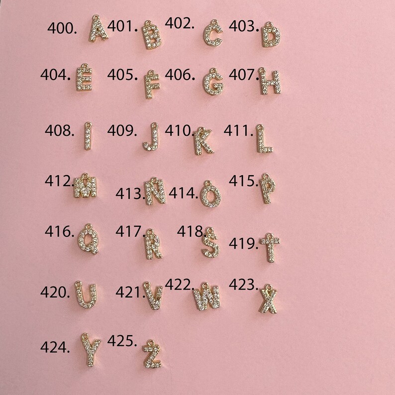 Puede incluir: Un conjunto de 26 abalorios de letras doradas, cada uno adornado con brillantes diamantes de imitaci&oacute;n transparentes. Los abalorios est&aacute;n dispuestos en orden alfab&eacute;tico, de la A a la Z, sobre un fondo rosa.