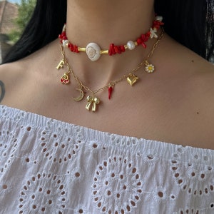 May include: Two gold necklaces with charms. The top necklace is made of red coral beads, white pearls, and a seashell. The bottom necklace has charms including a crescent moon, a bow, a heart, a chili pepper, a cherry, and a flower.