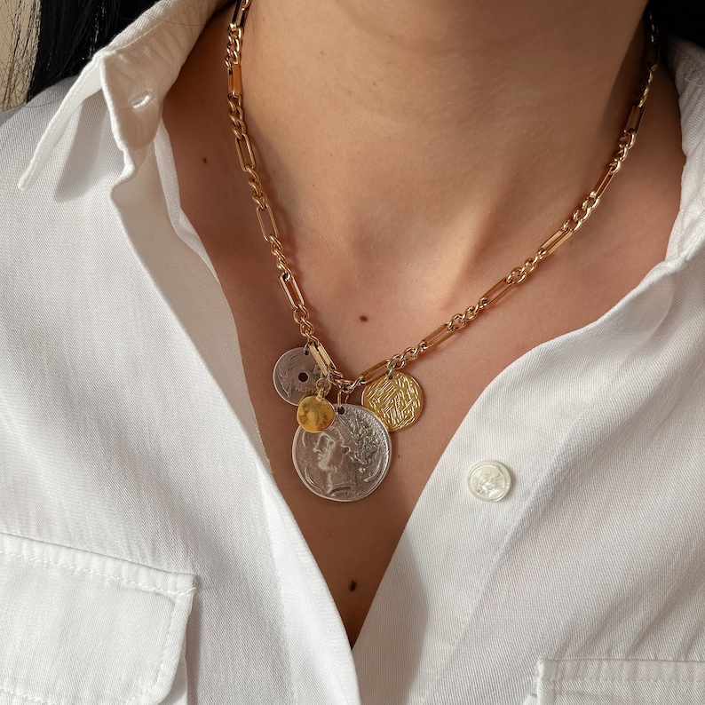Puede incluir: Un collar de cadena dorada con varios colgantes de monedas, incluyendo una moneda de plata con un retrato de perfil, una moneda de oro con escritura &aacute;rabe y una moneda de plata m&aacute;s peque&ntilde;a. El collar lo lleva una persona con una camisa blanca.