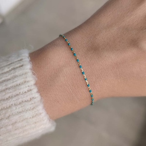 Puede incluir: Un delicado brazalete de cadena dorada con cuentas de color azul turquesa. El brazalete está en la muñeca de una persona.