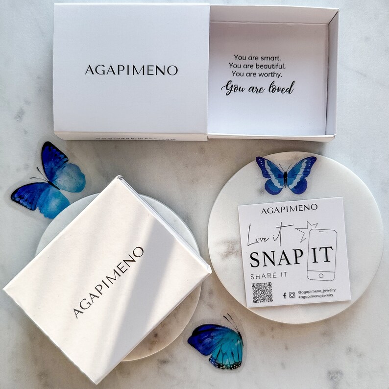 May include: A white box with the text "AGAPIME&Nu;&Omicron;" on the front. The box is open and inside is a white card with the text "You are smart. You are beautiful. You are worthy. You are loved."  The box is sitting on a white marble surface. There are two blue butterfly stickers on the surface.