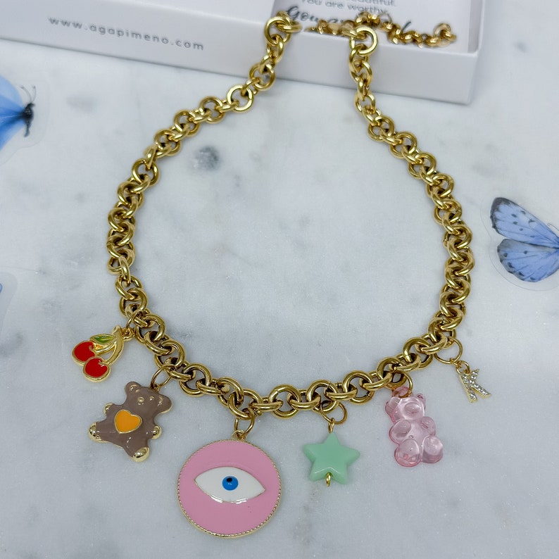 May include: Gold chain necklace with multiple charms, including a pink evil eye, a green star, a pink gummy bear, a brown teddy bear, and a red cherry. The necklace is on a white surface.