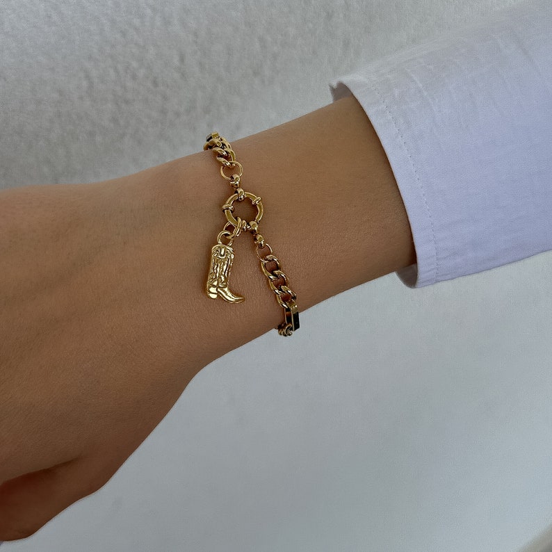 May include: A gold chain bracelet with a charm of a cowboy boot. The bracelet is on a person's wrist.