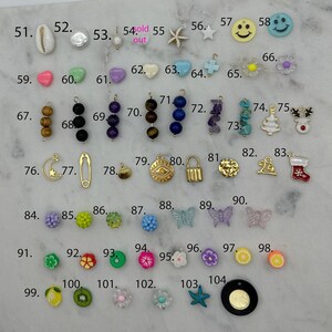 May include: Assortment of colourful charms and beads, including a seashell, a heart, a star, a smiley face, a flower, a Christmas tree, a stocking, a butterfly, a lemon, a kiwi, and a starfish. The charms are arranged in rows on a white surface.