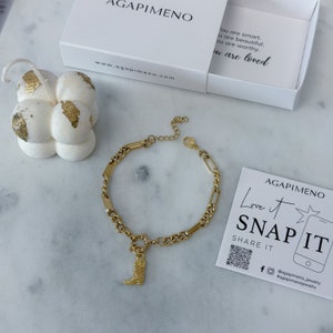 May include: Gold chain bracelet with a charm of a cowboy boot. The bracelet is on a white surface with a white box and a white candle with gold accents in the background. The text "Love it SNAP IT SHARE IT" is on a white card with a QR code and social media icons.