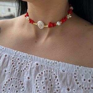 May include: A red coral and shell choker necklace with a gold accent. The necklace features a large white shell with a spiral design in the center.
