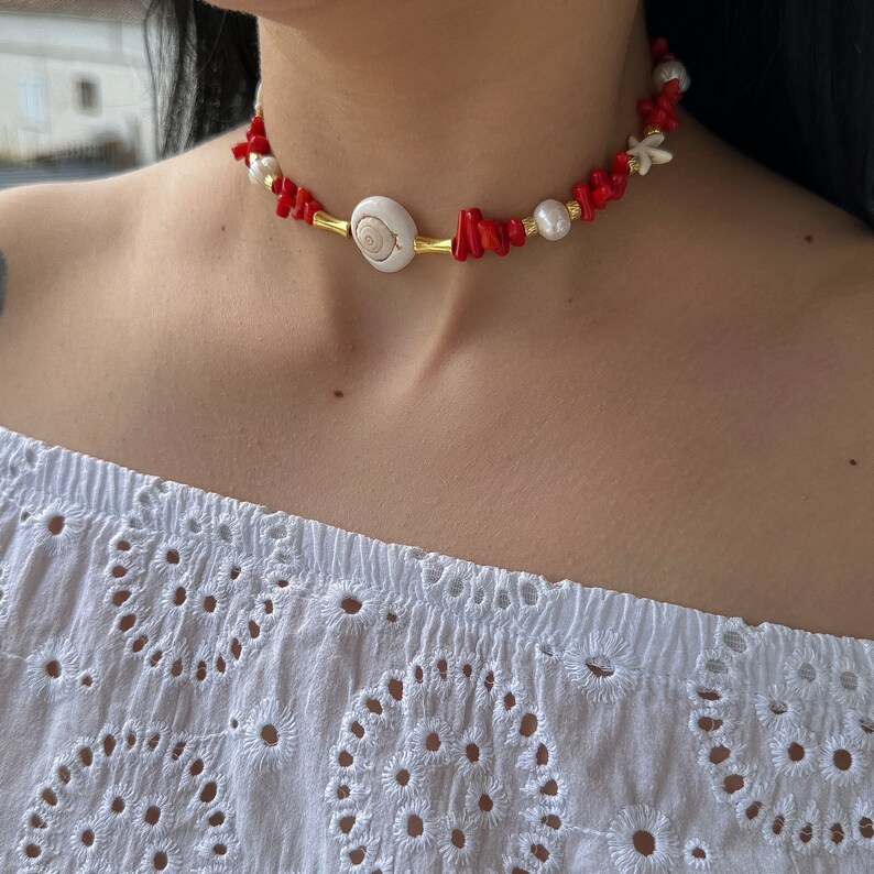 May include: A red coral and shell choker necklace with a gold accent. The necklace features a large white shell with a spiral design in the center.