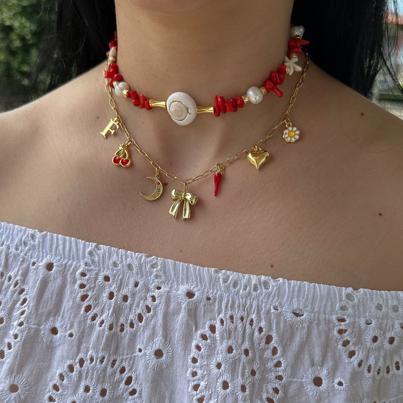 May include: A gold chain necklace with multiple charms, including a heart, a flower, a crescent moon, a cherry, a red pepper, and a bow. The necklace is layered over a red coral and white shell choker.