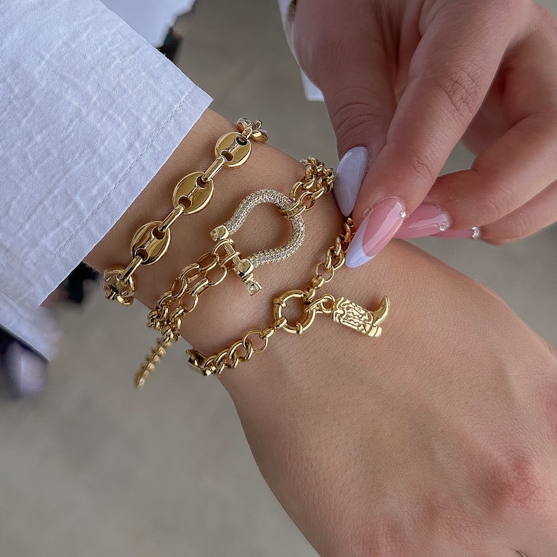 May include: Three gold bracelets with charms. The top bracelet is a chain with oval links. The middle bracelet has a horseshoe charm with clear rhinestones. The bottom bracelet has a boot charm.