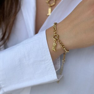 May include: A gold chain bracelet with a charm of a cowboy boot. The bracelet is on a person's wrist.