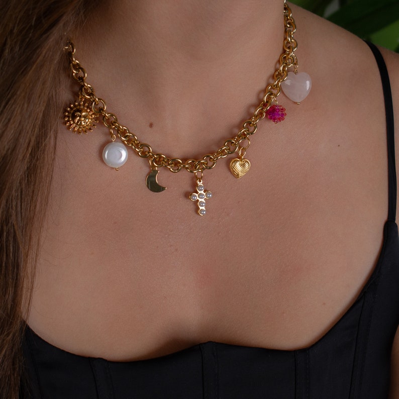 May include: A gold chain necklace with multiple charms, including a pearl, a crescent moon, a cross with small white stones, a heart, a pink heart-shaped stone, and a small gold flower.