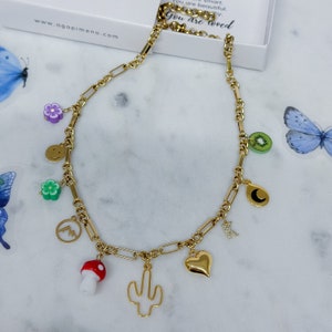 May include: A gold chain necklace with multiple charms, including a smiley face, a flower, a mountain, a mushroom, a cactus, a heart, a kiwi, a crescent moon, and a letter "N".