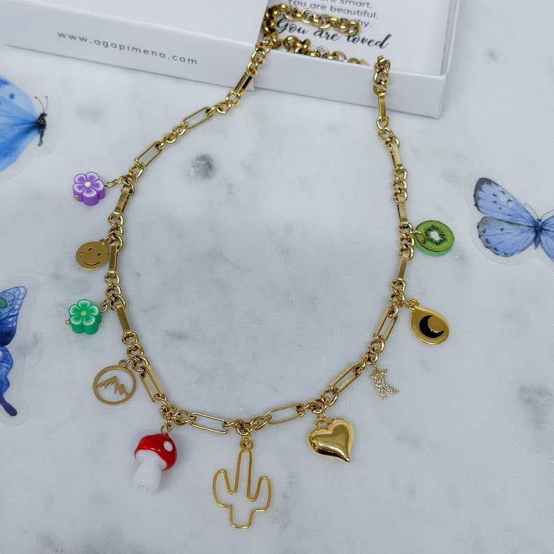May include: A gold chain necklace with multiple charms, including a smiley face, a flower, a mountain, a mushroom, a cactus, a heart, a kiwi, a crescent moon, and a letter "N".
