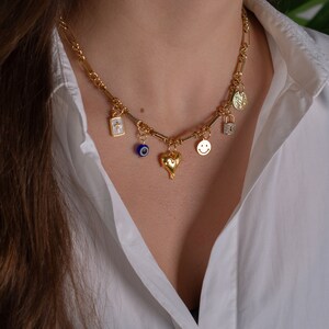 May include: A gold chain necklace with multiple charms, including a heart, a smiley face, a padlock, an evil eye, and a cross. The necklace is worn by a person with long brown hair.