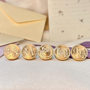 May include: Five gold-coloured wax seal stamps, each with a different letter from A to E. The stamps are circular with decorative designs. Envelopes and stationery are in the background.