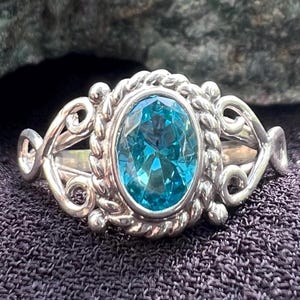 Peut inclure: Une bague en argent ornée d'une pierre précieuse ovale bleu clair. La pierre est sertie dans une lunette décorative en forme de corde. La bague présente des détails tourbillonnants ornés sur les côtés, créant un design élégant et accrocheur.