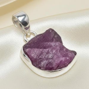 May include: A raw, uncut, purple gemstone pendant with a silver-coloured bail. The pendant has a rough, textured surface and is set in a polished silver bezel. The background is a soft, off-white satin fabric.